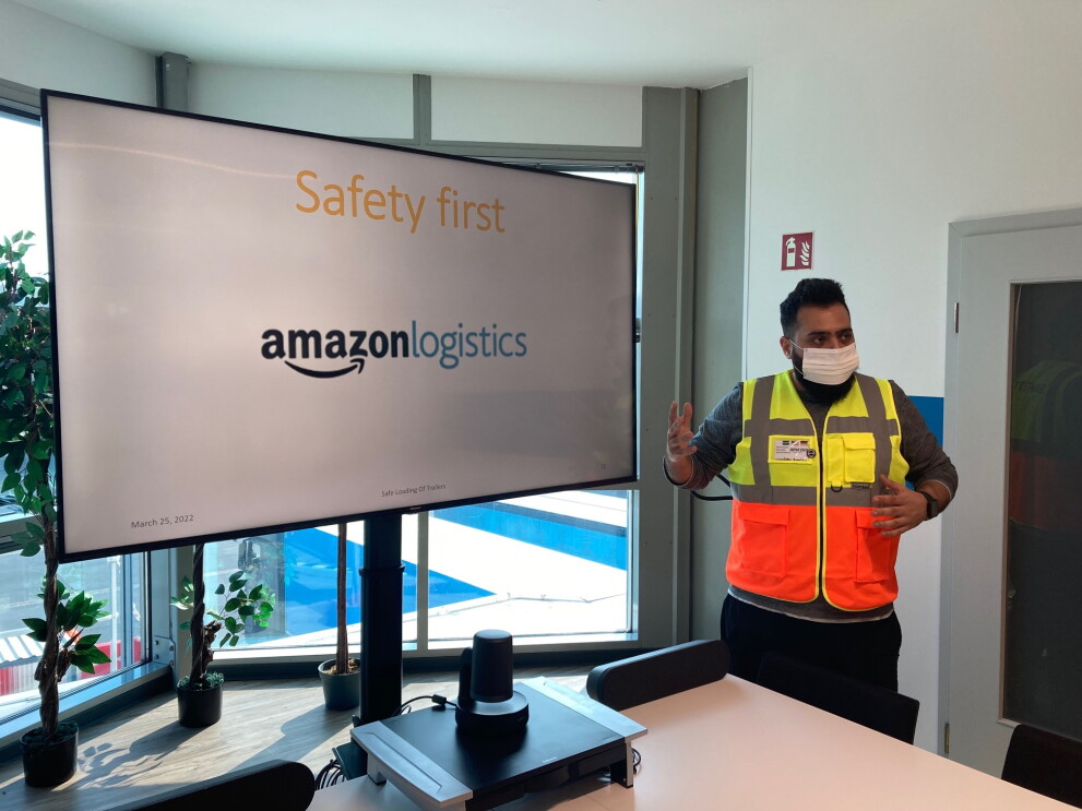 Spezialist für Arbeitssicherheit bei Amazon Mustafa Almujahed bei einer Schulung zu Thema Safety first. Er steht mit einer gelb-orangenen Weste und Maske bekleidet vor einem Schildschirm mit der Aufschrift "Safety first. Amazon logistics".