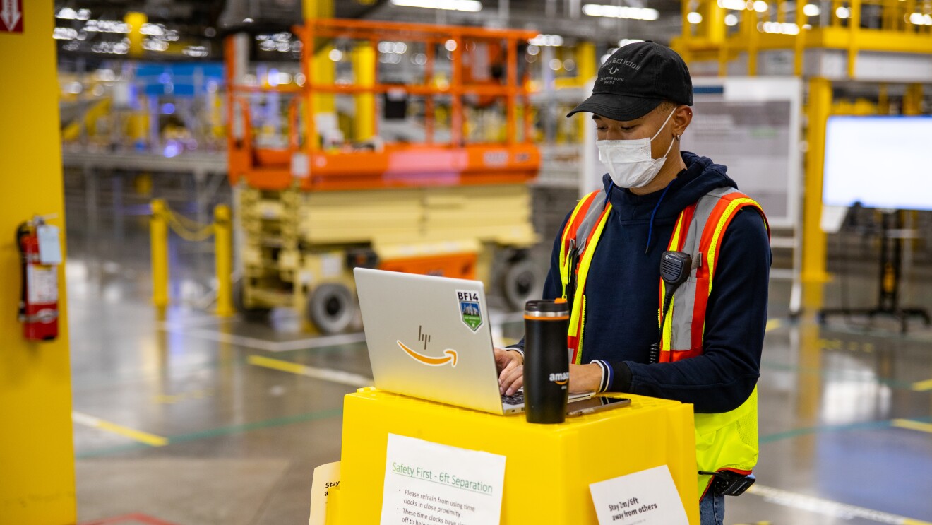 在亚马逊物流中心,一名男子站在一堆黄色的提包旁边,在电脑上打字。