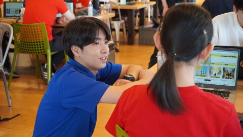 机の上にノートパソコンを広げて座る赤いTシャツの女性の後ろ姿。青いTシャツの男性がかがんで、机に両腕を載せて話しかけている。背景には同じ机が複数並び、他の参加者が見える。