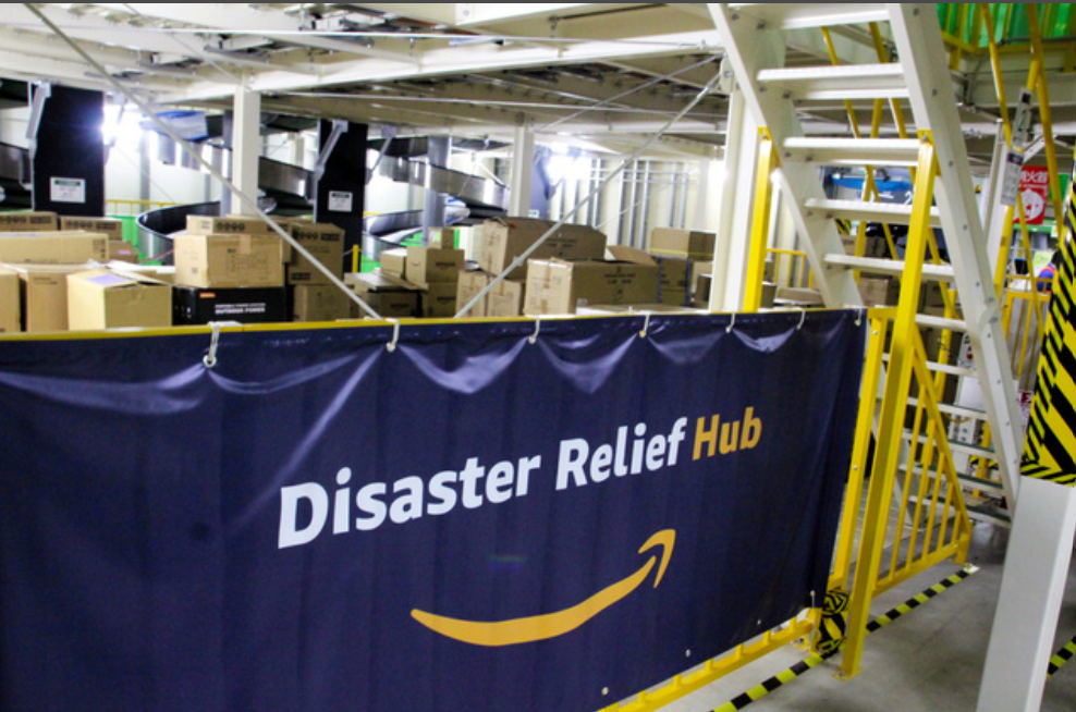 フルフィルメントセンター内に  Disaster Relief Hubを書かれた横断幕。その向こうに段ボールに入った物資が見える