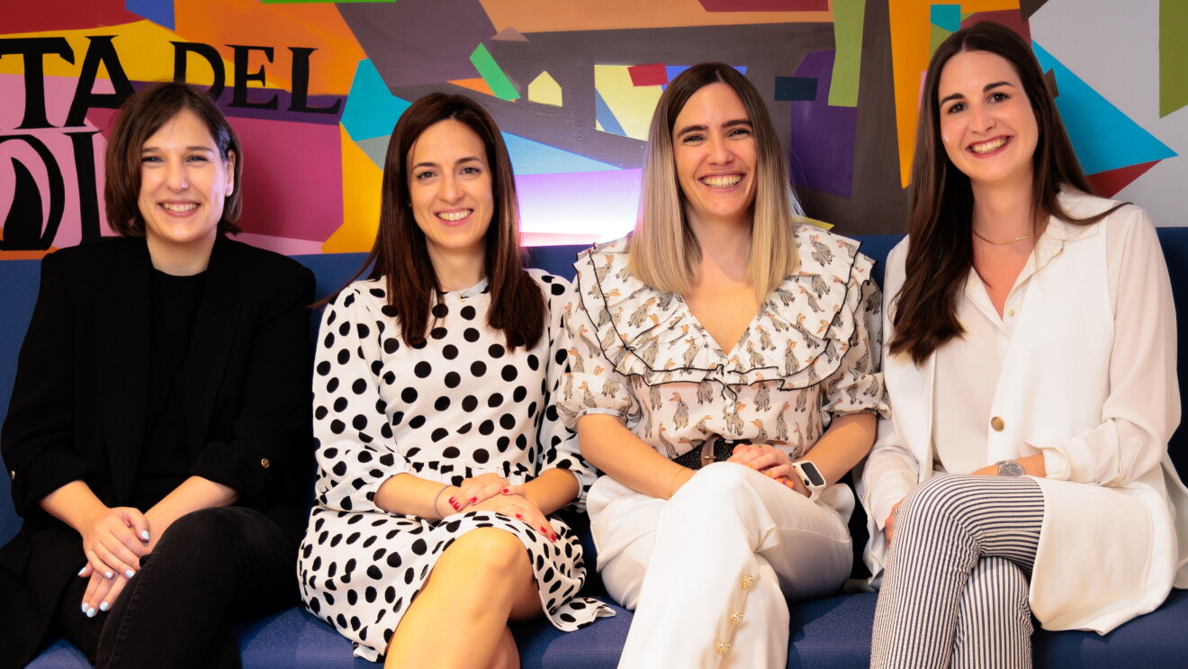 Sentadas en un sofá de color azul y con un fondo pintado de muchos colores vivos y las letras en negro de Puerta del Sol, están sentadas cuatro mujeres con la pierna izquierda cruzada. De izquierda a derecha: María Alonso vestida con un traje pantalón de color negro, con un jersey negro también debajo de la americana. Lleva unas sandalias marrones con dos tirantes. Al lado está Alejandra Ossa tiene el pelo negro largo hasta el hombro. Lleva un vestido blanco de lunares negros de manga larga y largo hasta la rodilla.  Lleva unas sandalias de color de oro de plata. Lara Fajardo es la cuarta de la foto. Va con una camisa blanca estampada, con el pelo hasta el hombro con mechas negras y decoloradas hasta el rubio en las puntas. Con pantalones blancos y unas sandalias negras de plataforma de color de mimbre de color marrón y negro. Y, finalmente, Miriam Domínguez-Adame Aguilera con el pelo liso negro hasta el pecho. Unas sandalias negras, unos pantalones rayados de color negro y blanco, una camisa y chaleco de color blanco.