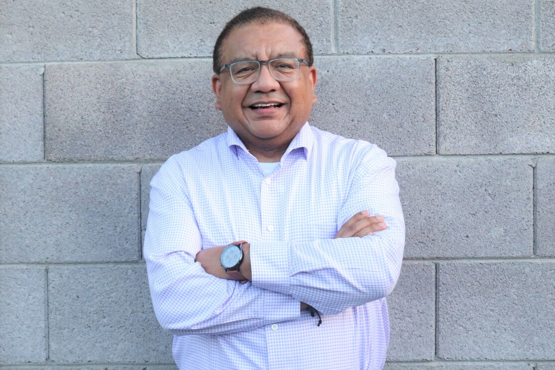 An image of a man smiling for a photo while folding his arms while standing in front of a cement brick wall.