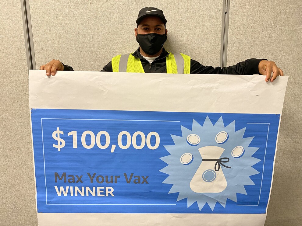 An image of a man holding a large check made out for $100,000 that says "Max Your Vax Winner." He is wearing a mask and a yellow safety vest. 