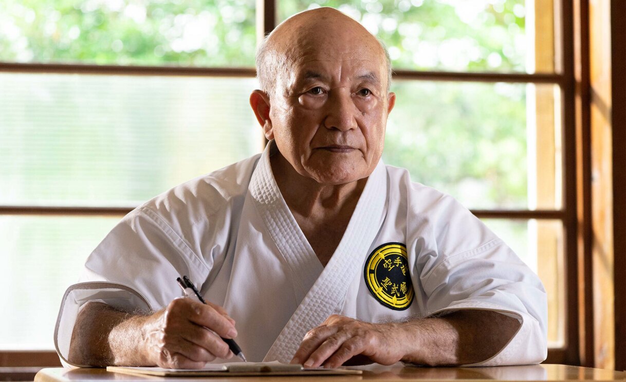 An image of Zenbo Shimabukuro, director of Shorin-ryu Seibukan, sitting on a desk and holding a pen in his hand. 