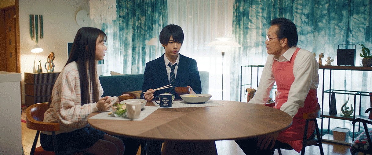 青と白のカーテンがかかった室内。丸いテーブルに向って3人の人物が座っている。左側の2人だけ食事をしていて、右側のピンクのエプロンの男性は座っている。