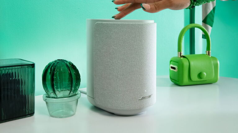 Hand hovering over white fabric smart speaker surrounded by green glass cactus decoration and green handbag on turquoise background