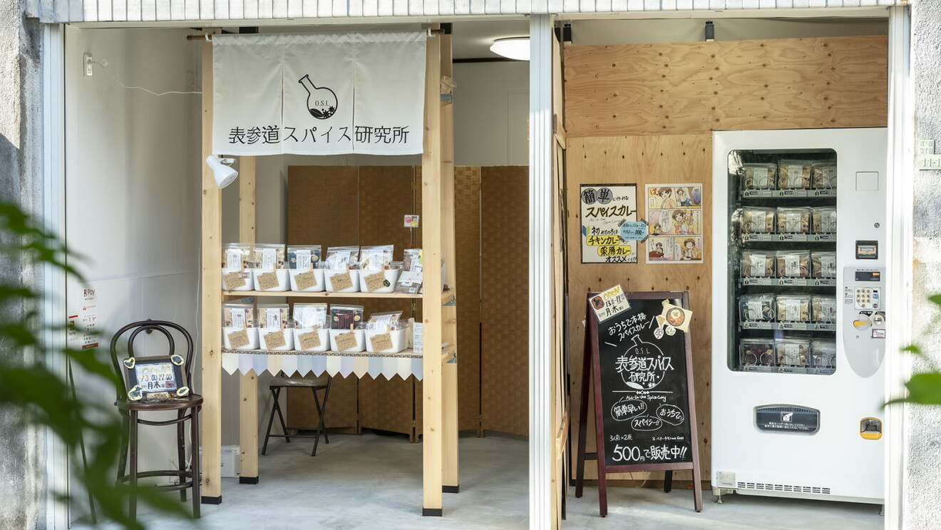 白や木の壁の店舗に、木製棚と自動販売機が設定されている。棚の上部には「表参道スパイス研究所」と書かれたのれん。棚にも自動販売機にもスパイス商品が並ぶ。