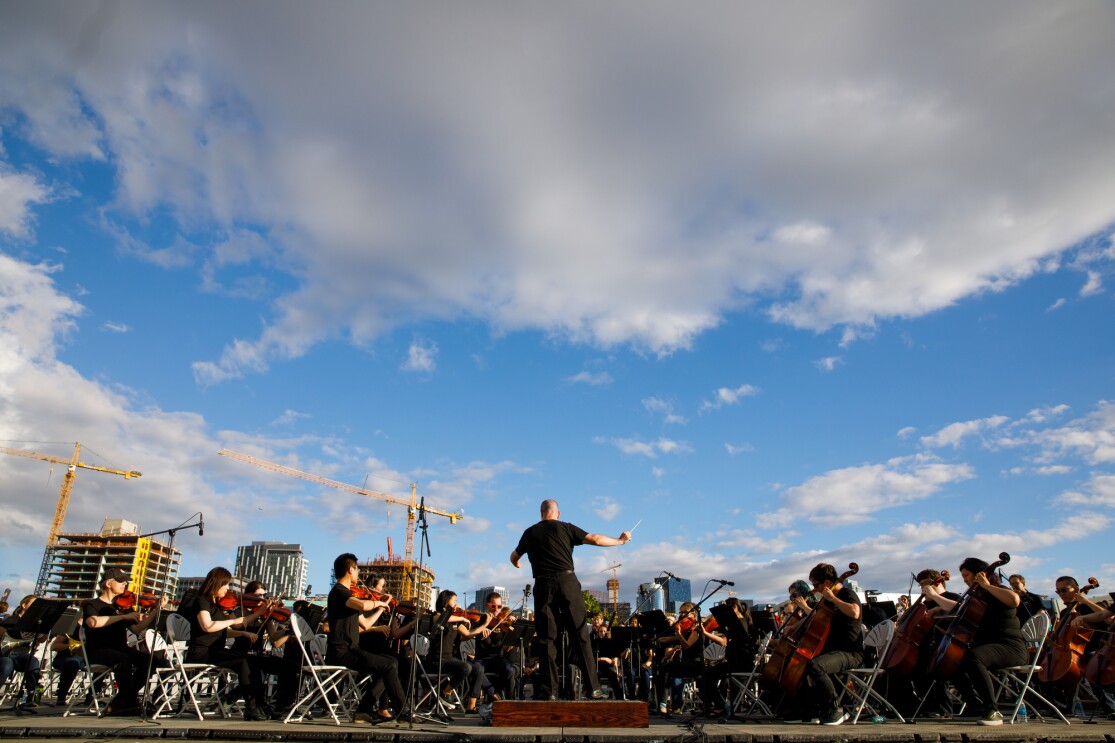 In front of a clear, blue sky, with a few buildings and cranes visible, the Amazon Symphony Orchestra performs at the closing ceremony. In this image, a conductor is seen on a platform, surrounded by orchestra members with string-based instruments.