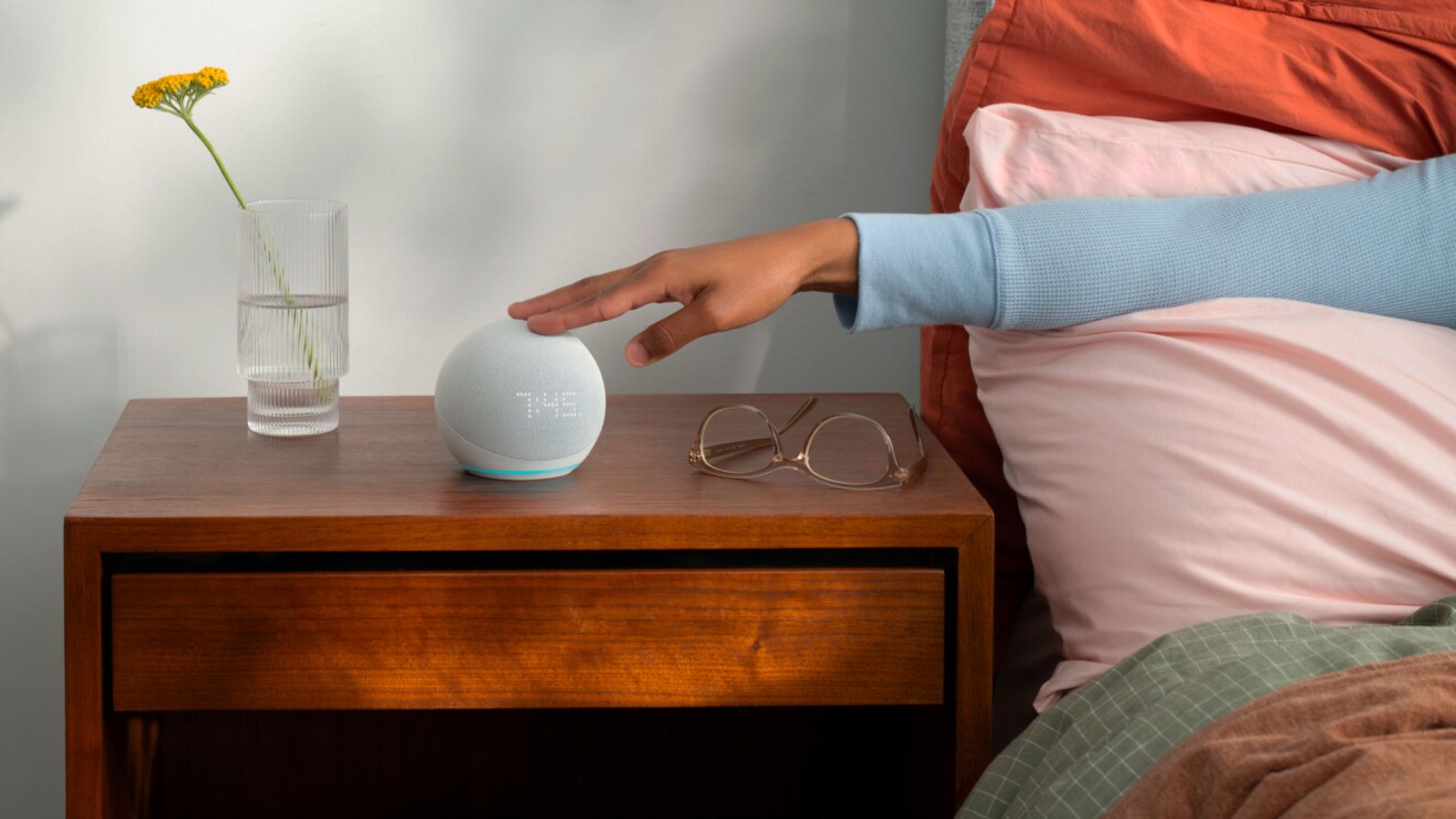 An image of a wooden night stand and with an arm touching an Echo device from a bed. There's a pair of glasses and a vase with a flower next to the device. 