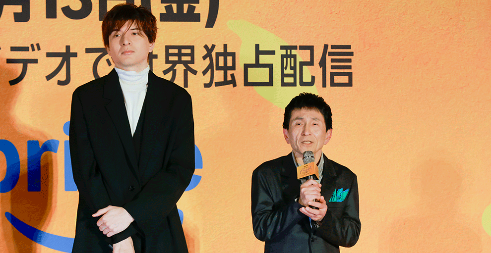 オレンジ色の壁の前、オレンジのステージの上で並ぶ2人の人物。右側のグレーの上下の男性がマイクを手に持って話している。