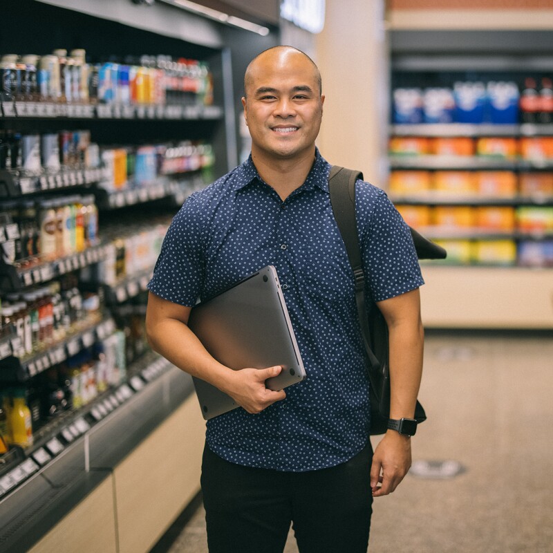 A man holding a laptop computer in one arm smiles at the camera from within an Amazon Fresh grocery store.