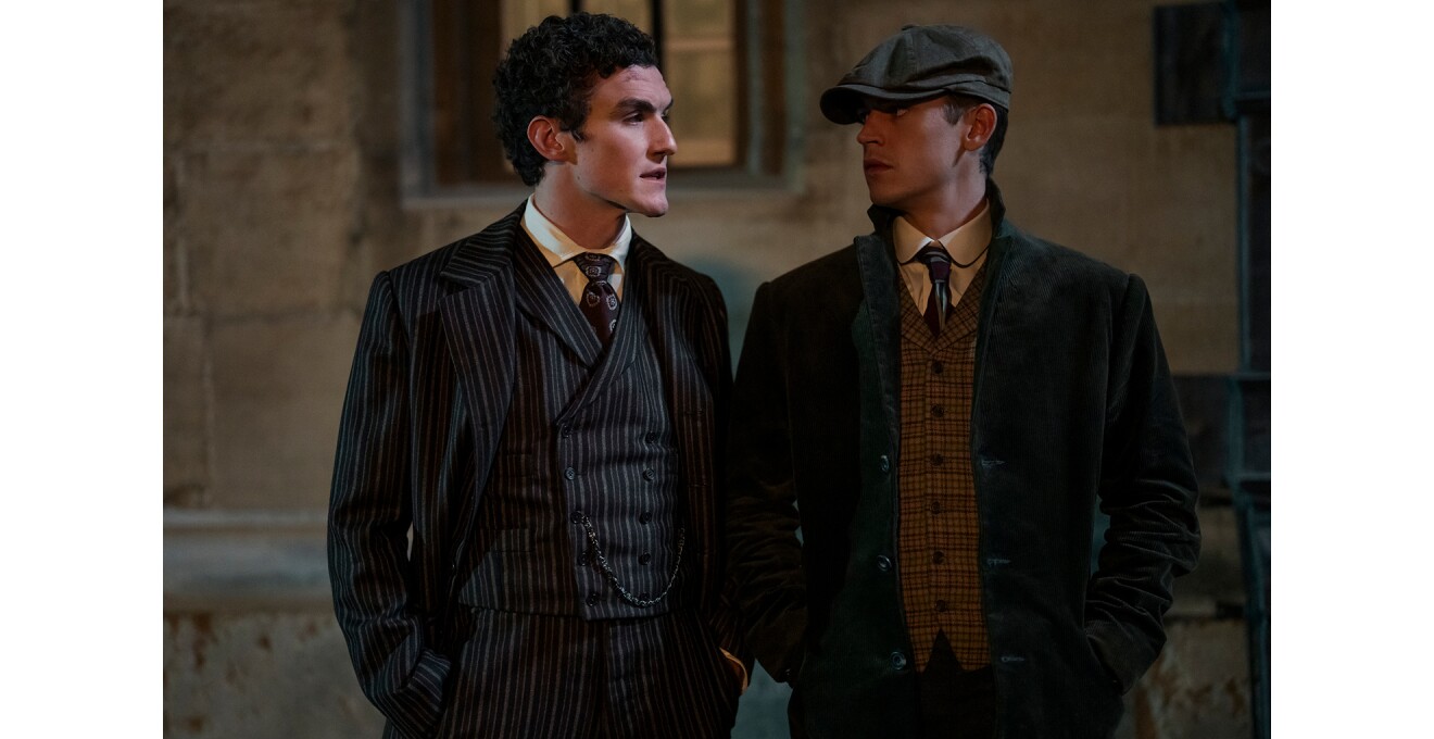 薄暗い屋外。家屋の壁を背に、三つ揃いスーツの男性とジャケットにベスト、ハンチング棒の男性が並んで立って見つめ合っている。