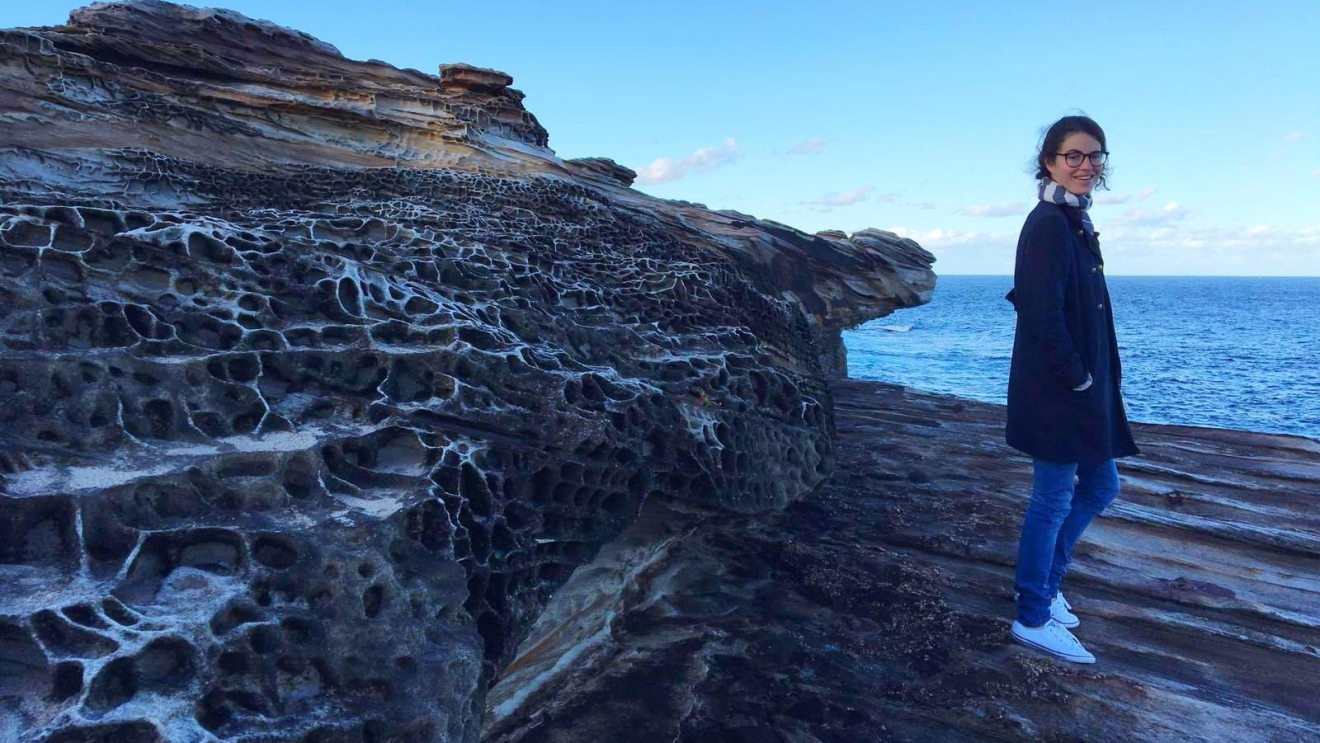 An image of a woman standing on a rocky beach in a coat and scarf smiling for the photo.