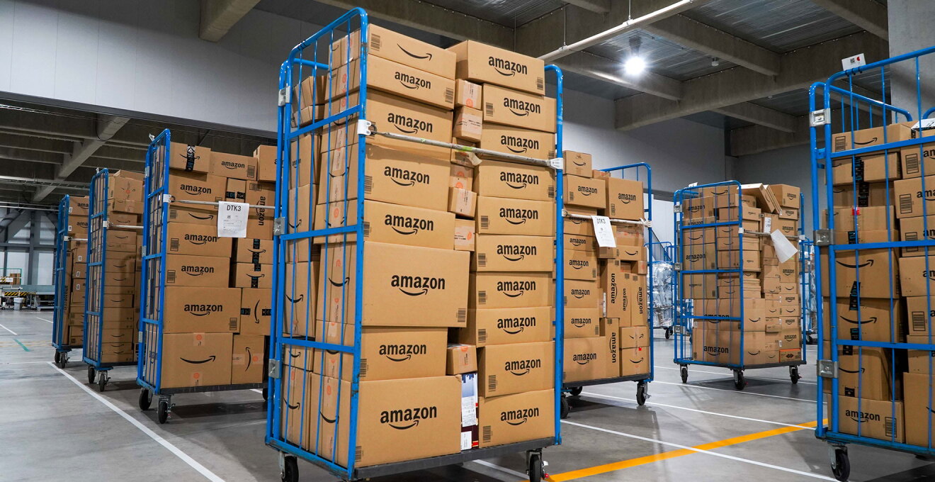 グレーの物流倉庫内、Amazonロゴ入りダンボールを満載した青い台車がたくさん並んでいる。床には白とオレンジの線が引かれている