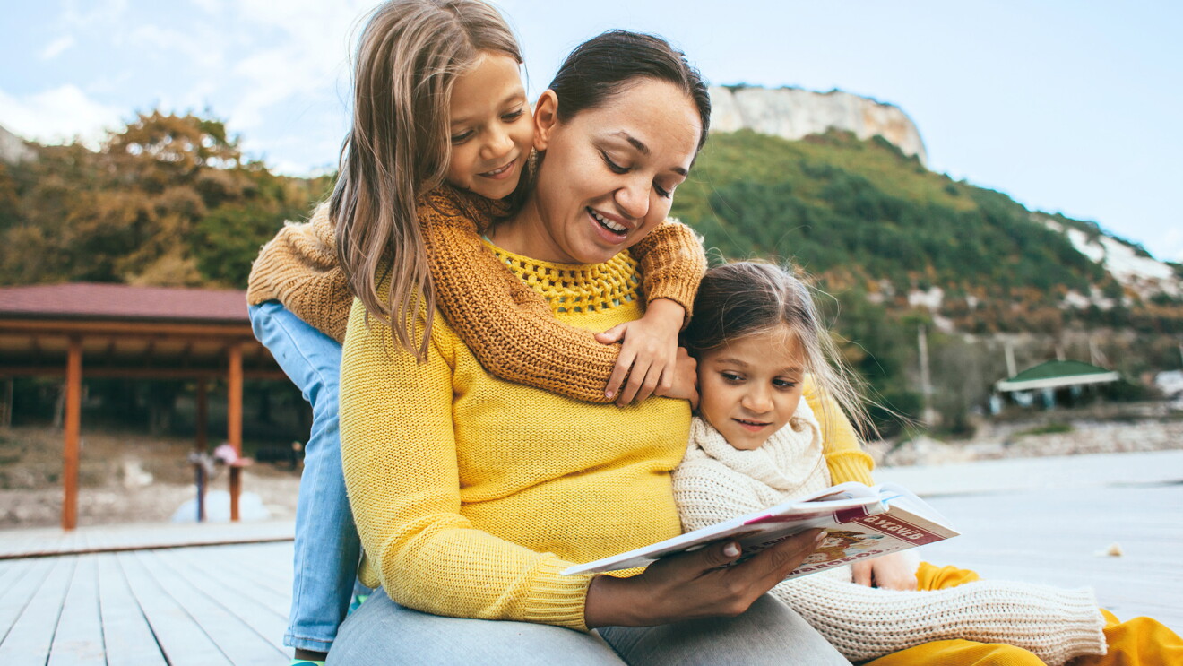 A woman wearing a sweater sits on a dock with two small girls, reading a story book to them. 