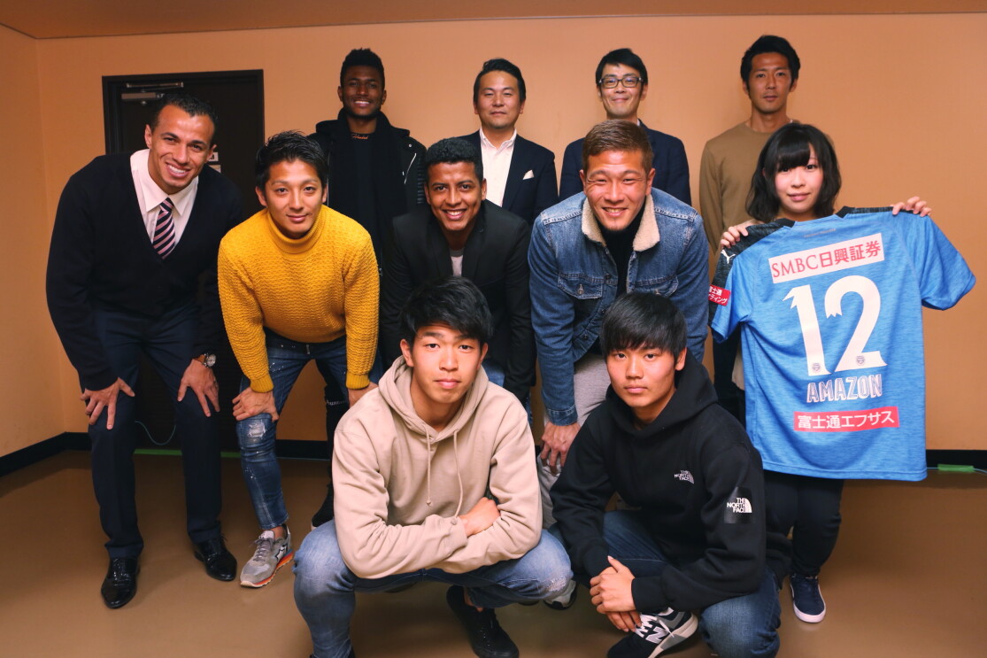 ベージュの壁の室内で集合写真を撮る11人の人物。前列はしゃがみ、中列は膝に手をついて姿勢を低くし、後列は直立。