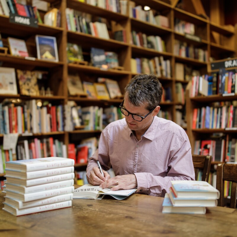 An author sits at a table and signs his book in a bookstore, surrounded by shelves of other book titles.