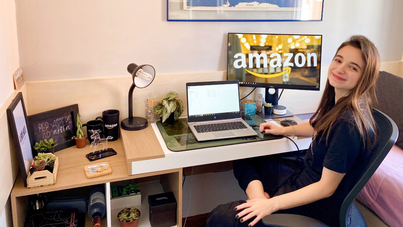 A woman sits at a desk with a laptop and a monitor showing the Amazon logo.