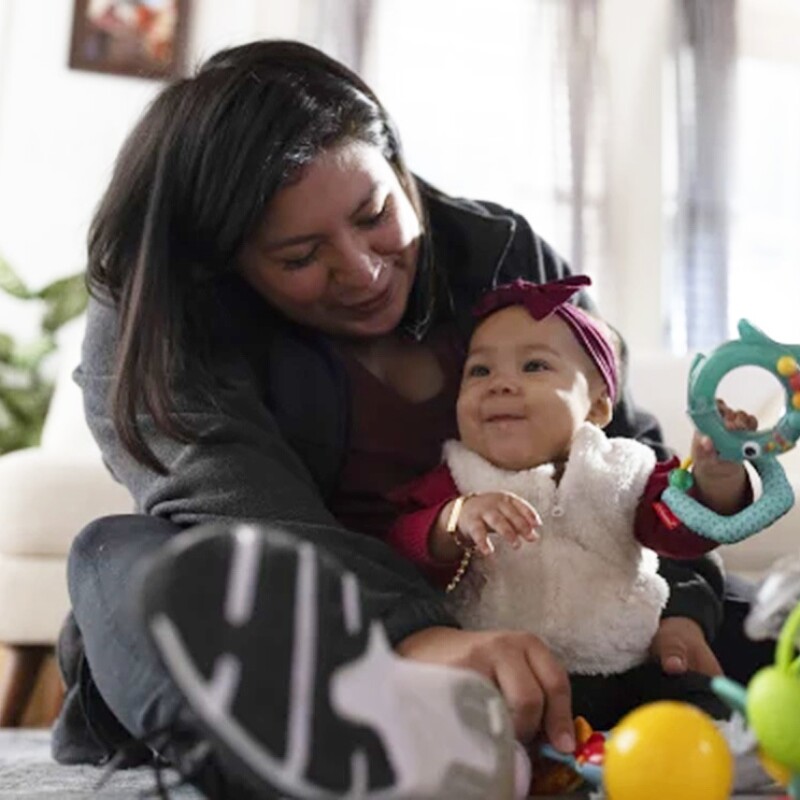 A woman sits with a baby surrounded by toys, while a man looks on.