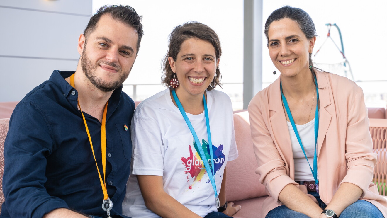Simone Fortunini, Coia Pons y Eva Ahijado, miembros de glamazon, en las oficinas corporativas de Amazon en Madrid.  Los tres están sentados, miran a cámara y sonríen. Simone viste con una camisa azul, Coia con la camiseta de glamazon y Eva con una camiseta blanca y una americana de color salmón.
