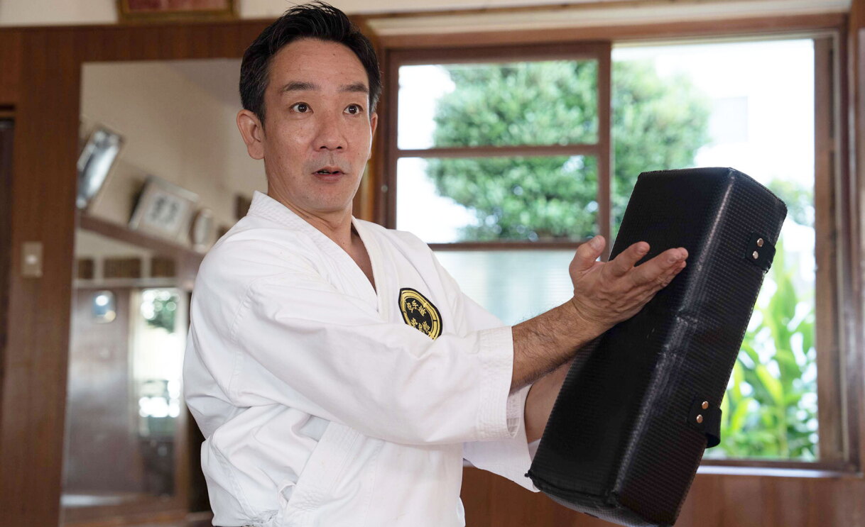 An image of a man teaching karate in Shorin-ryu Seibukan, one of Okinawa's traditional karate schools. He is holding a back block in one hand and pointing to it with the other.