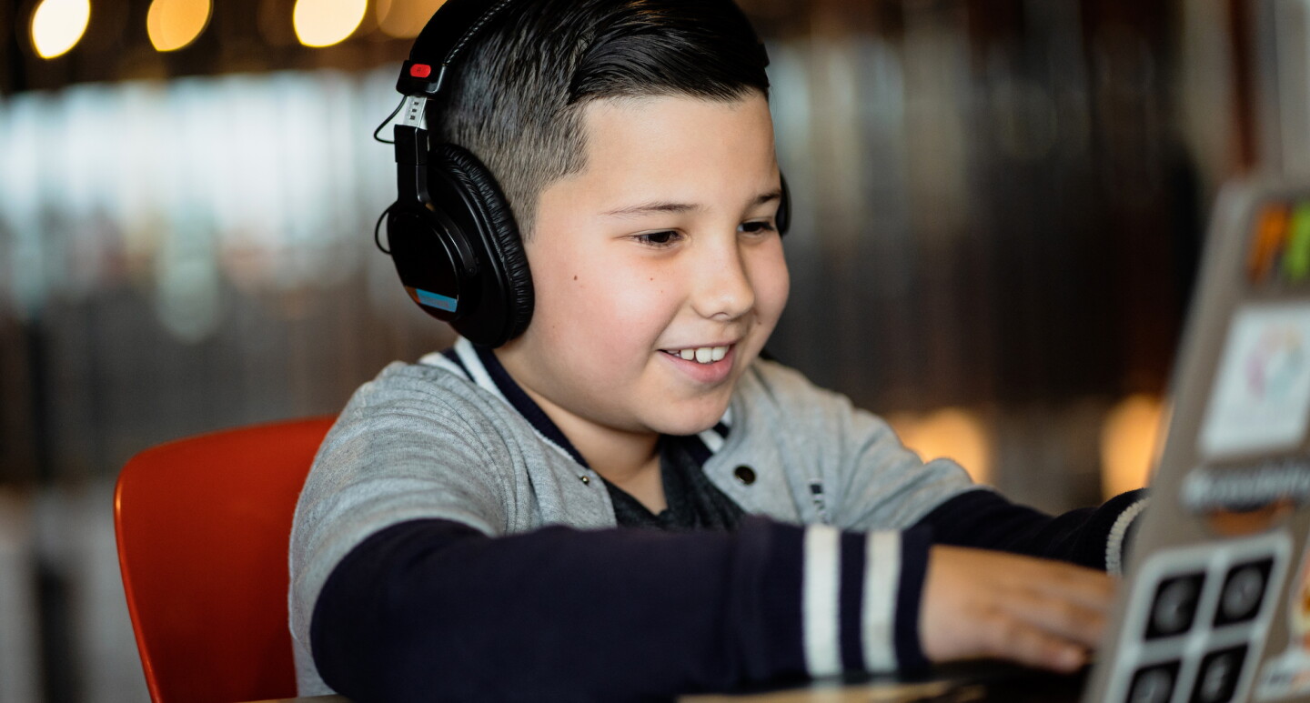A boy wearing headphones works on a laptop at a coding camp while smiling.