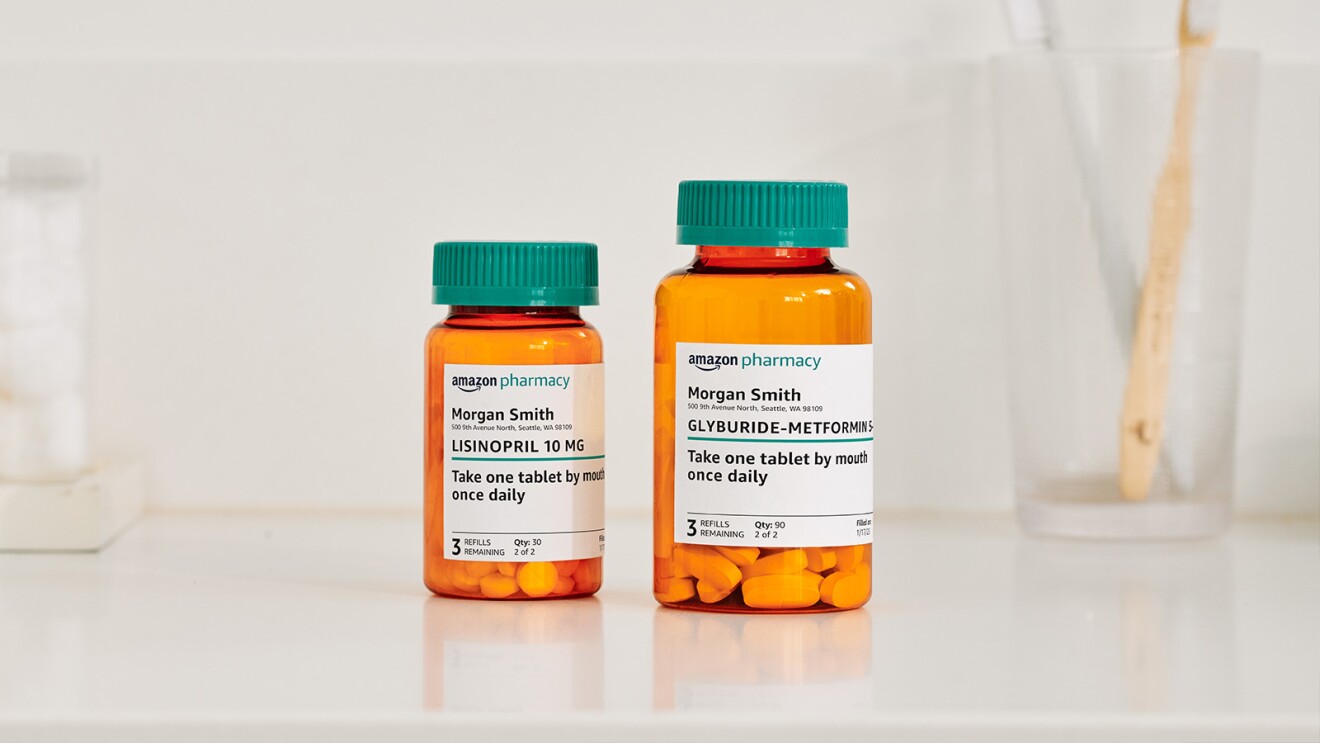 Two Amazon Pharmacy pill bottles sit on a white bathroom countertop next to a glass with two toothbrushes in it.