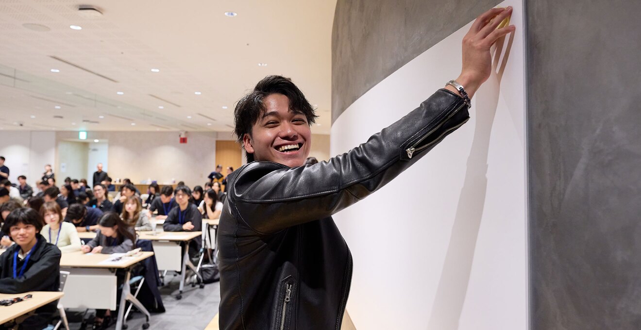 壁に貼られたホワイトボードに手を伸ばして触れる笑顔の若者。ホワイトボードにはまだ記入も貼り付けもされていない。