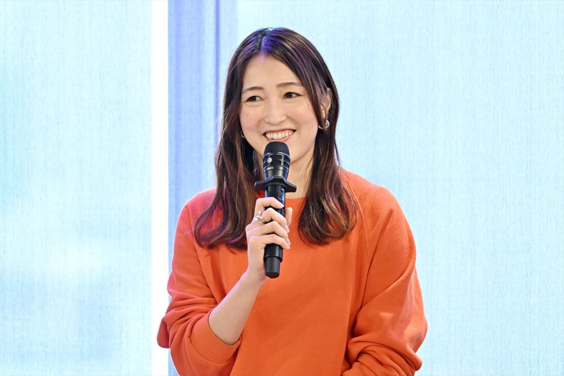 オレンジ色のセーターを着ている女性がマイクを持って微笑んでいる