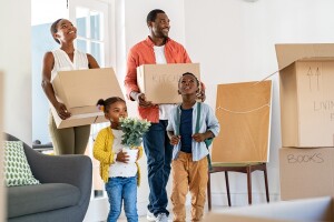 A photo of a family standing in the living room of a house. They are holding moving boxes and there are moving boxes placed in the living room.