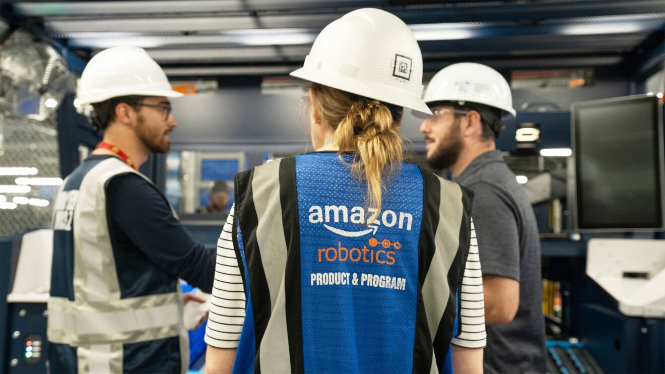 An image of three employees in hard hats, one employee is in a vest that reads Amazon Robotics Product and Program