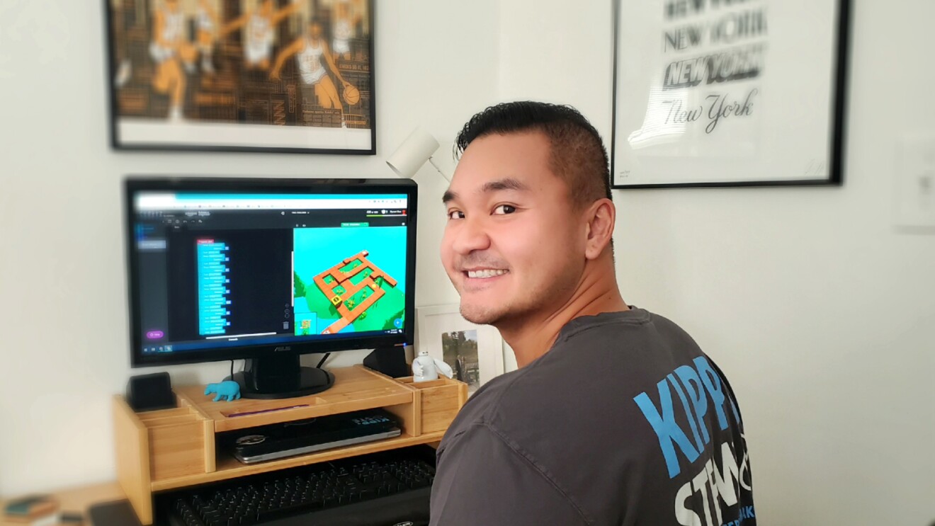 A man sitting at a desk with a computer on it turns around and smiles toward the camera.