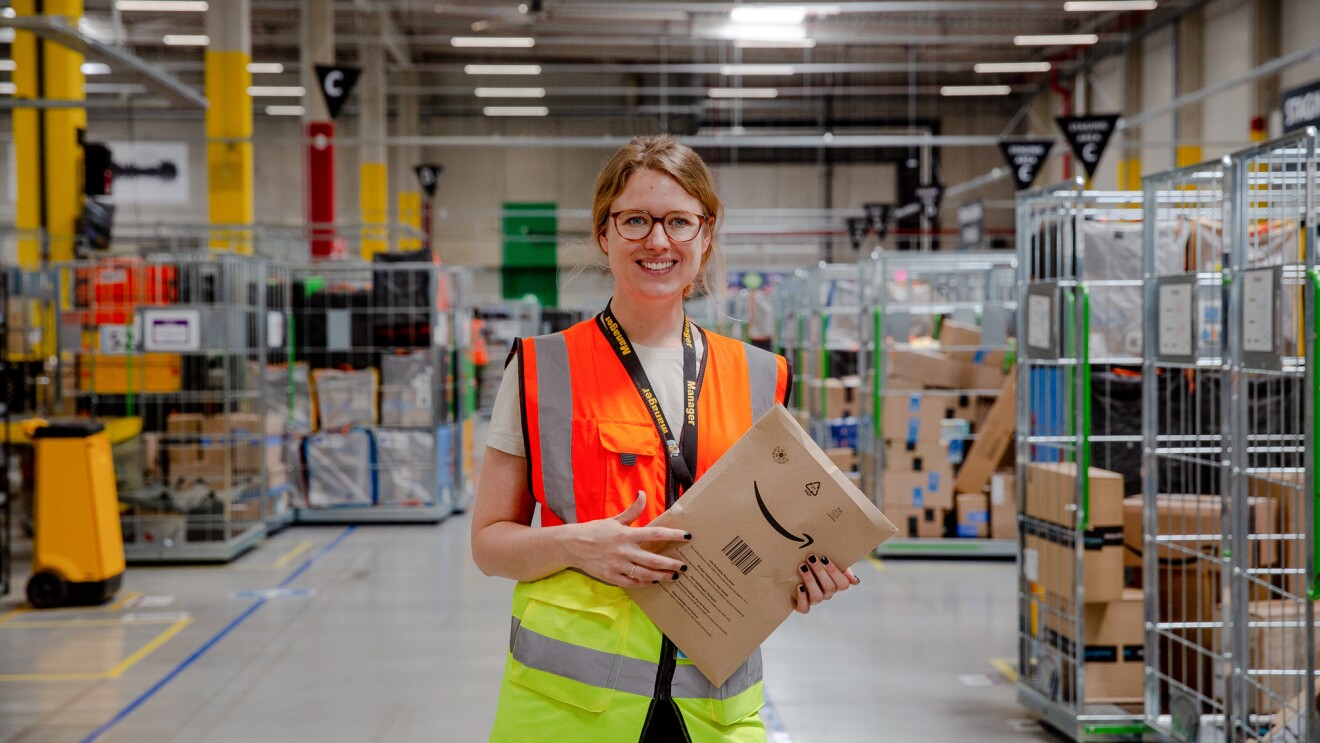 Regionalleiterin Miriam Enzi trägt eine gelb-orangene Weste und hält ein Amazon-Versandpäckchen in der Hand, während sie in einem Logistikzentrum von Amazon in Österreich steht.