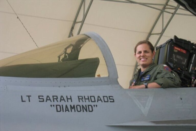 An image of Sarah Rhoads in the cockpit of a jet plane. She is smiling and wearing her full pilot's uniform. There is text on the side of the plane that reads "Lt. Sarah Rhoads 'Diamond'"