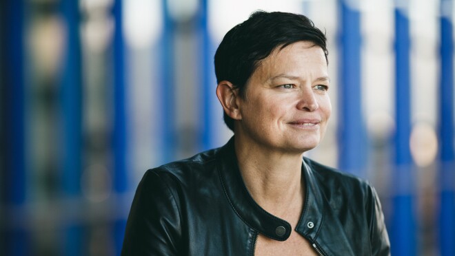 一头黑发,亚马逊的救灾贝蒂娜斯蒂克斯,身着一件黑色皮夹克看起来是正确的和微笑。有一个蓝色背景。