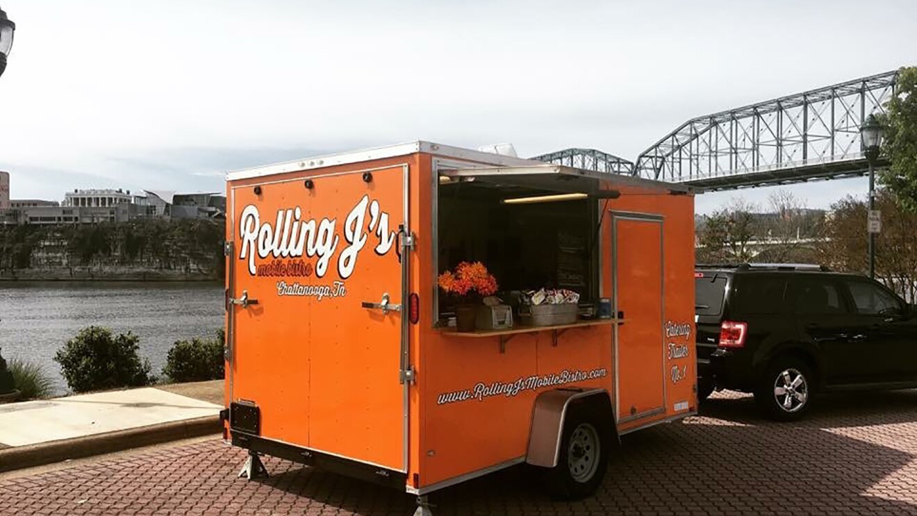 An orange food truck parked in front of the Tennessee River. The food truck has "Rolling J's" branding on it, and is parked on a street made of pavers.