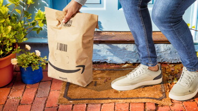 En el portal de una casa aparecen las piernas y la mano de una persona recogiendo una bolsa de papel de Amazon. 