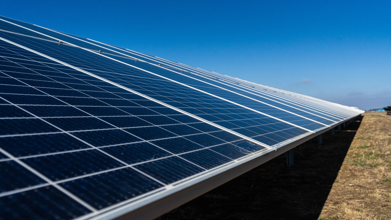 Primera plano de placas solares. El suelo es de tiera con hierbas de color verde y marrón. El cielo es azul.