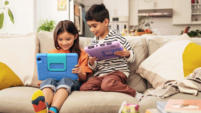 An image of two kids playing with their tablets sitting on a sofa.