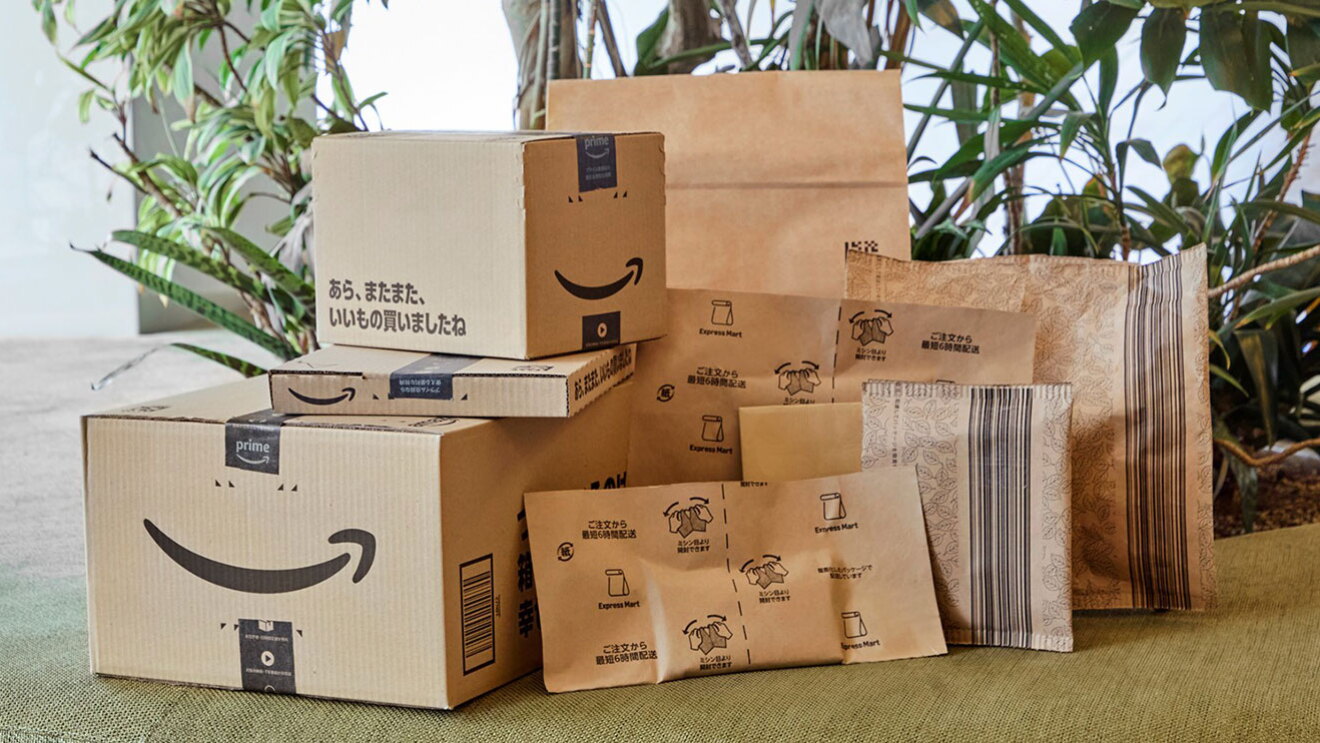 Amazonロゴが入った梱包資材が積まれている。大小のロゴ入り箱や平たい箱、ミシン目が入った大小の紙袋など、全部で9種類ある。背後に観葉植物。