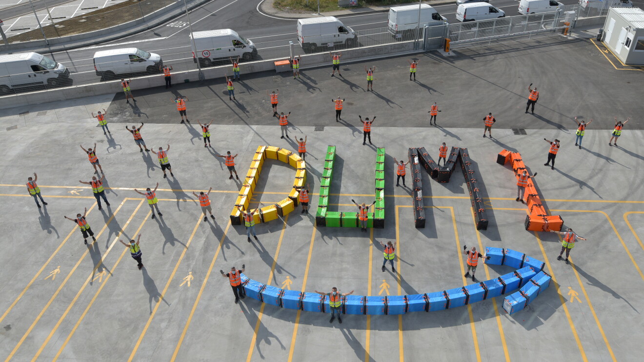 Foto aerea di un gruppo di dipendendi di un magazzino Amazon in Umbria posa con le braccia aperte e rivolte verso l'alto. A terra, alcuni sacchi colorati formano lo smile Amazon in azzurro e la scritta DUM1