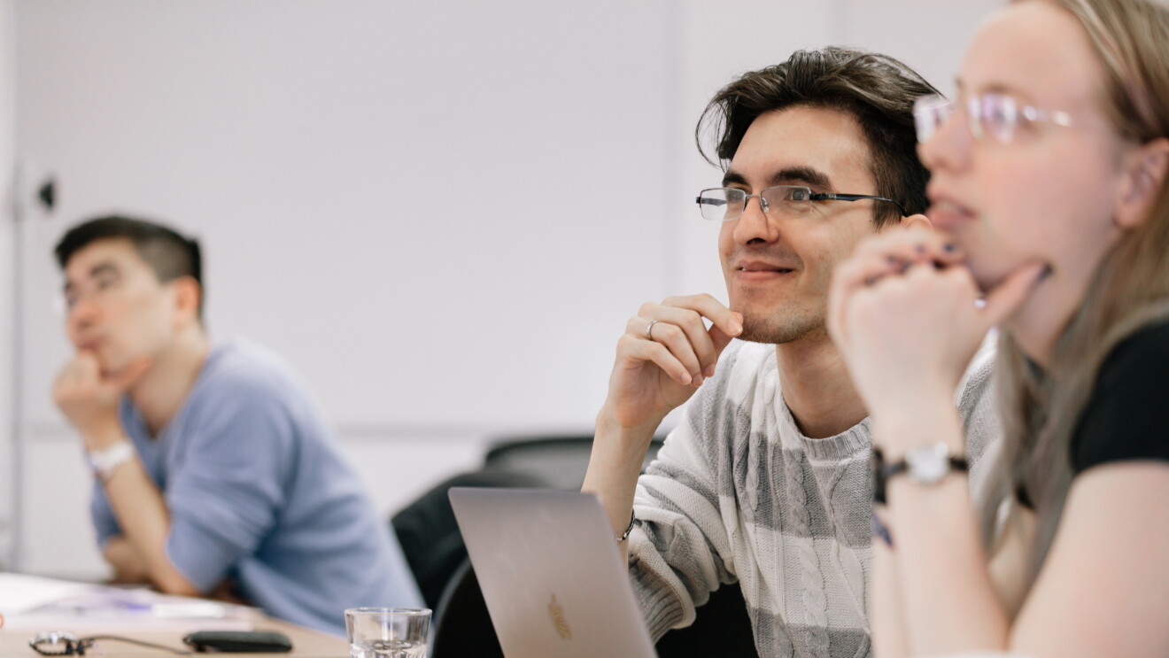 Students at the University of Oxford sitting in a lecture, they are sitting at their desks with their laptops, notes and drinks on the table. 