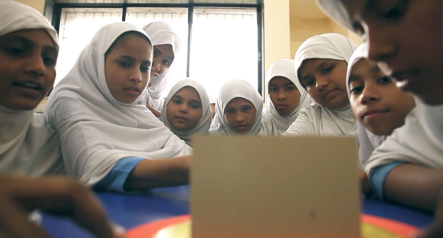 female schoolchildren sit gathered around and facing a small computer screen.