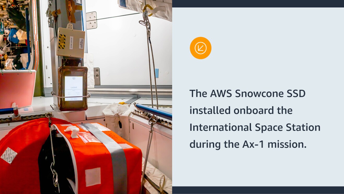 An image of an orange box with a lit up screen sitting on a white surface in a space ship. Text on the right of the image says "The AWS Snowcone SSD installed onboard the International Space Station during the Ax-1 mission."