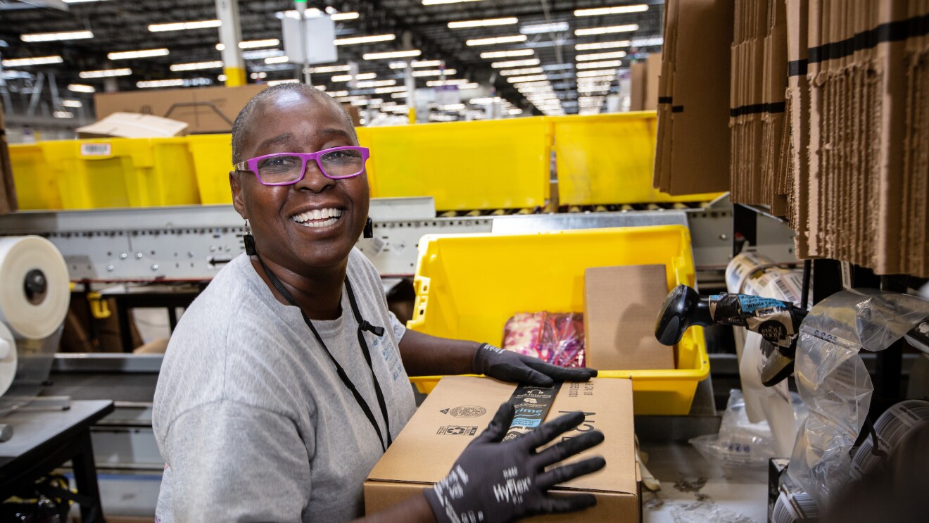 An Amazon Fulfillment Center employee smiles at the camera as she prepares a package for shipment.