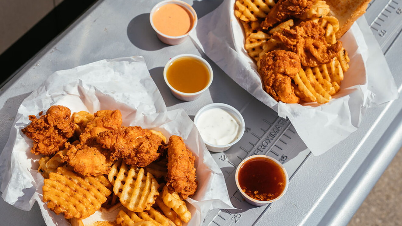 A basket of waffle fries, Texas toast, and chicken tenders, on ledge of a food truck window. To the right are four dipping sauces, and another basket of food. 