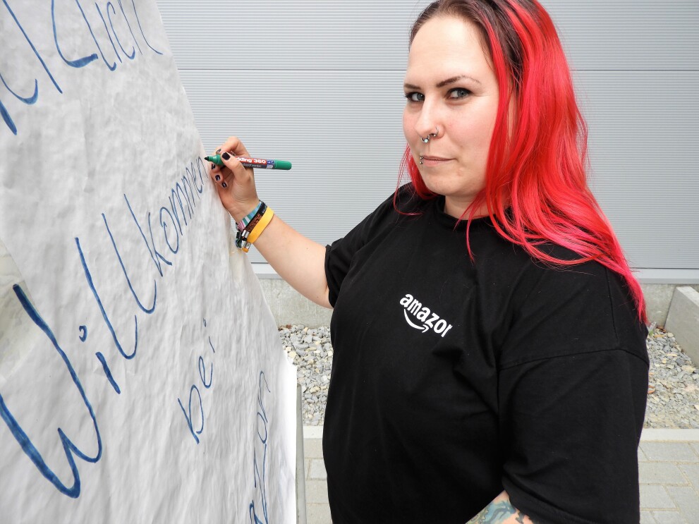 Eine rothaarige Frau mit langen Haaren und schwarzem T-Shirt mit der Aufschrift "Amazon"  schreibt auf ein Flip-Chart "HERZLICH WILLKOMMEN": Das ist Bentje.