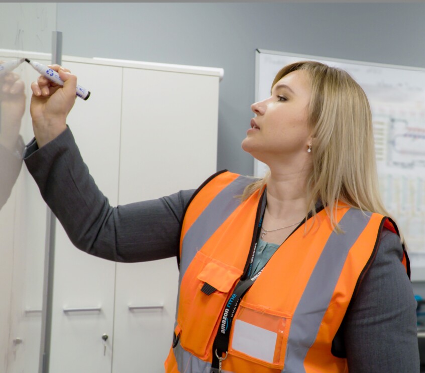 Eine blonde Frau mit orangener Sicherheitsweste über dem grauen Hosenanzug steht in einer Logistikhalle. Ihr Blick ist auf ein Blatt gerichtet, das sie in der Hand hält.