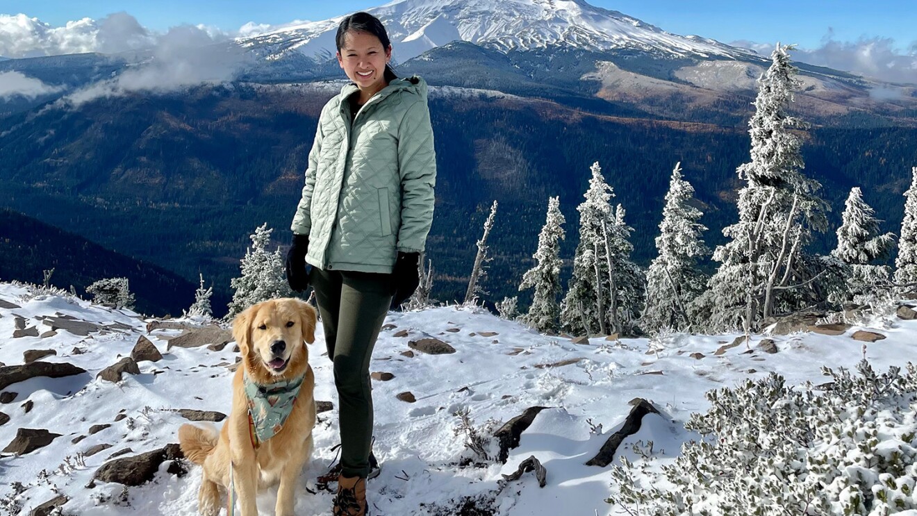 一个女人和她的狗站在一起拍照。背景是一座大山和白雪皑皑的冬季景观,蓝天。