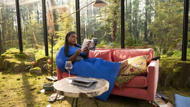 Uzo Aduba坐在家里的马车上,有大窗户可以看到外面的绿色景观,绿色的草像地毯一样穿过房子。她穿着一件与绿色形成鲜明对比的亮蓝色连衣裙。